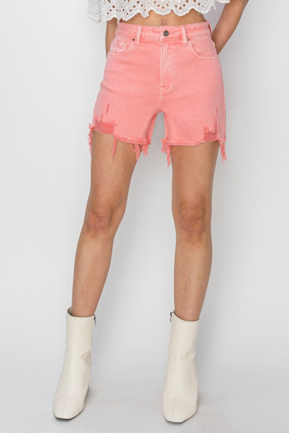 Sporting a stylish look with RISEN High Rise Distressed Denim Shorts in frayed pink and a white lace top, someone stands against a plain backdrop. The distressed details of the denim add an edgy flair to this chic outfit.