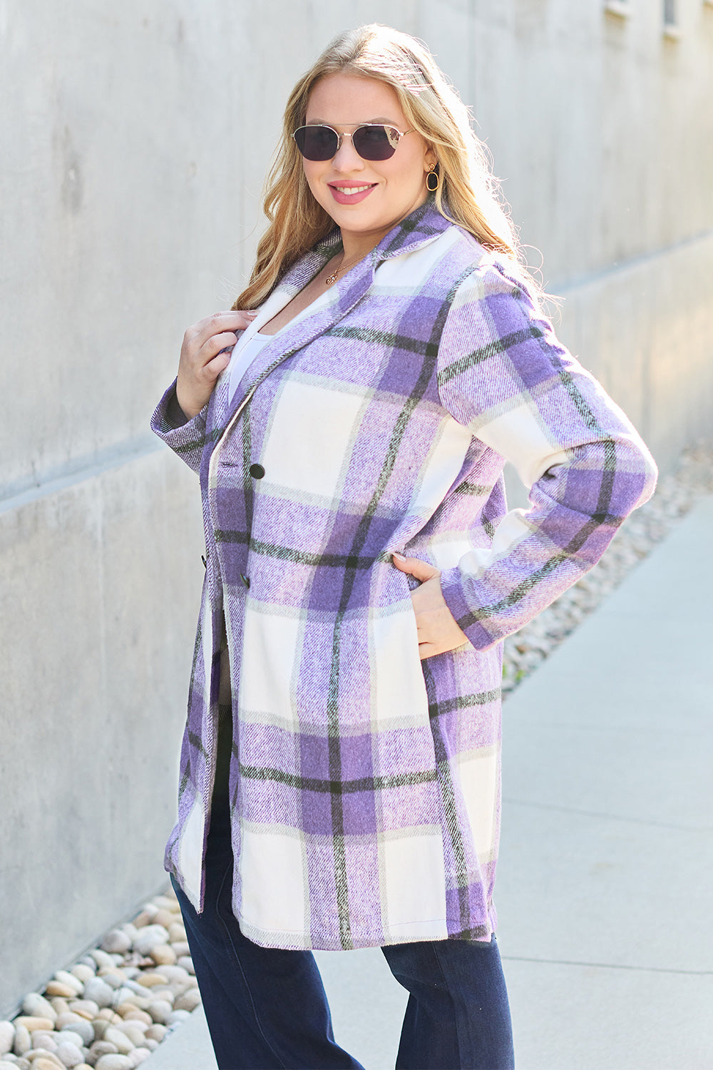 A woman stands on a sidewalk wearing the Double Take Full Size Plaid Button Up Lapel Collar Coat in pink and white over a white top and blue jeans, with her right hand lightly touching her hair. The polyester fabric of the coat makes it durable and perfect for an easy machine wash cold.