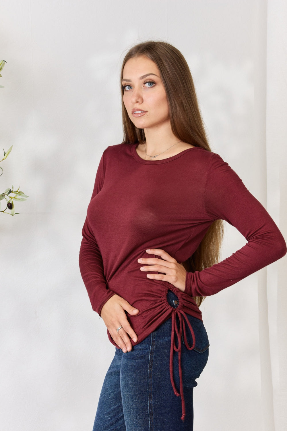 A woman stands in front of a plain background and a small tree, wearing the Culture Code Full Size Drawstring Round Neck Long Sleeve Top in maroon and blue jeans.