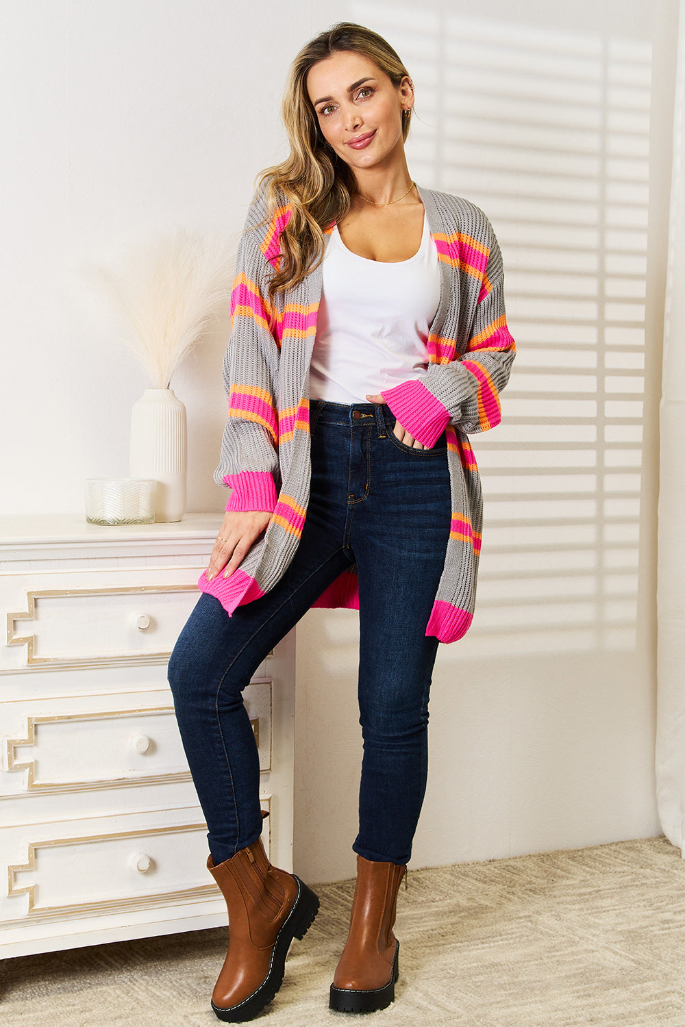 A woman stands against a white wall, smiling with one hand on her hip. She wears the Woven Right Ribbed Long Sleeve Cardigan, featuring pink and orange stripes, over a white top and dark jeans—an excellent choice for versatile layering.