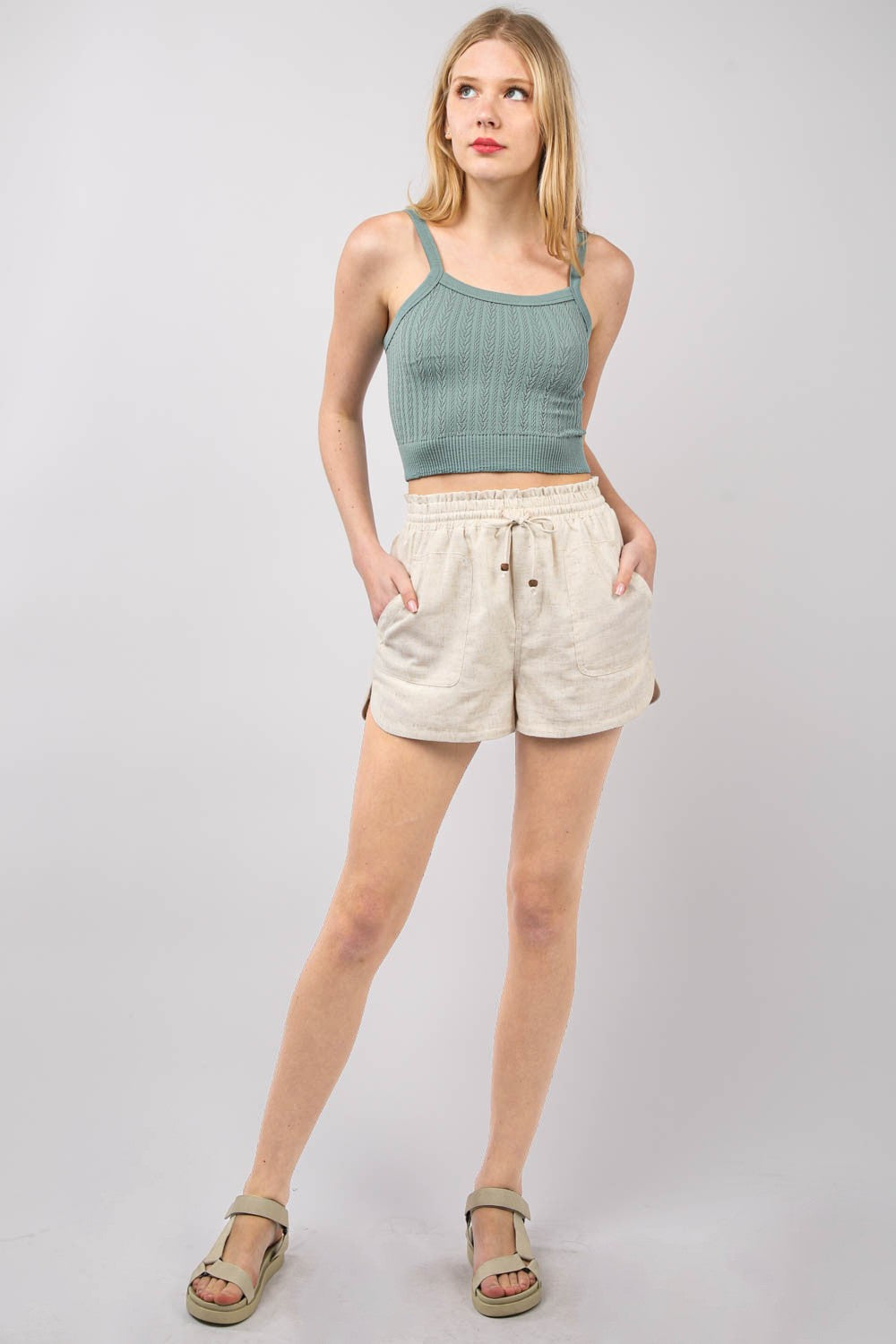 A person wearing VERY J Drawstring Elastic Waist Linen Shorts in beige and a green top. The image focuses on the midsection, with the eco-friendly shorts featuring front pockets and a slight texture.