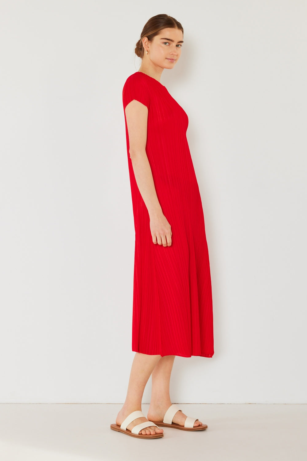 A person stands against a light wall, wearing an elegant Marina West Swim Pleated Cap Sleeve A-Line Dress in black and white sandals.