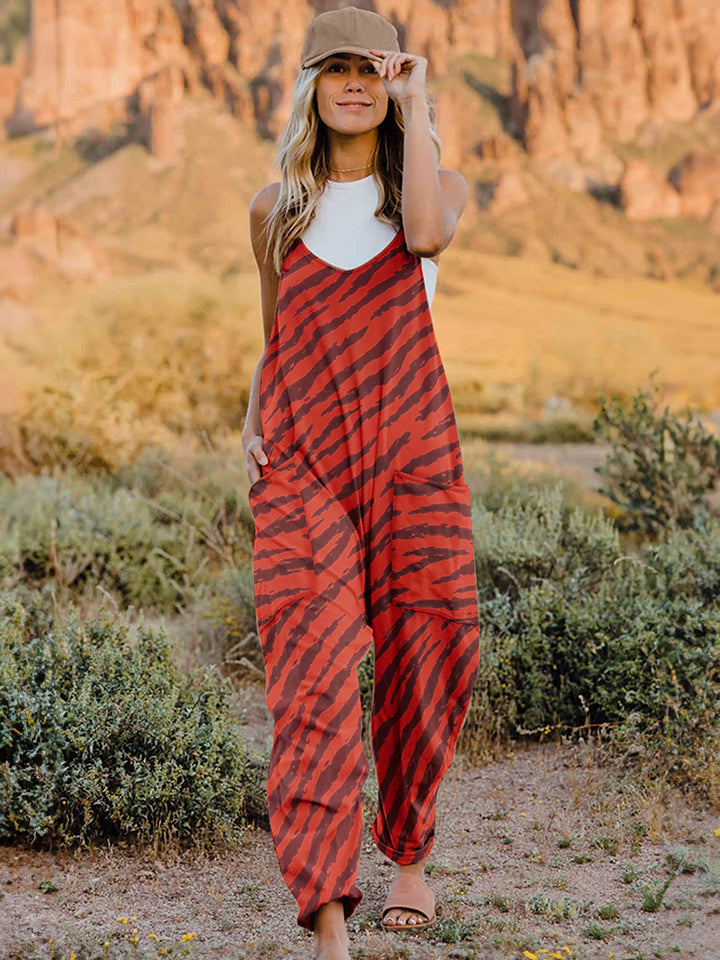 A person wearing a casual white V-neck tank top and a Full Size Printed V-Neck Sleeveless Jumpsuit with a brown animal print zebra-stripe pattern stands outdoors with one hand on their hat and the other in their pocket, set against a rocky landscape backdrop.