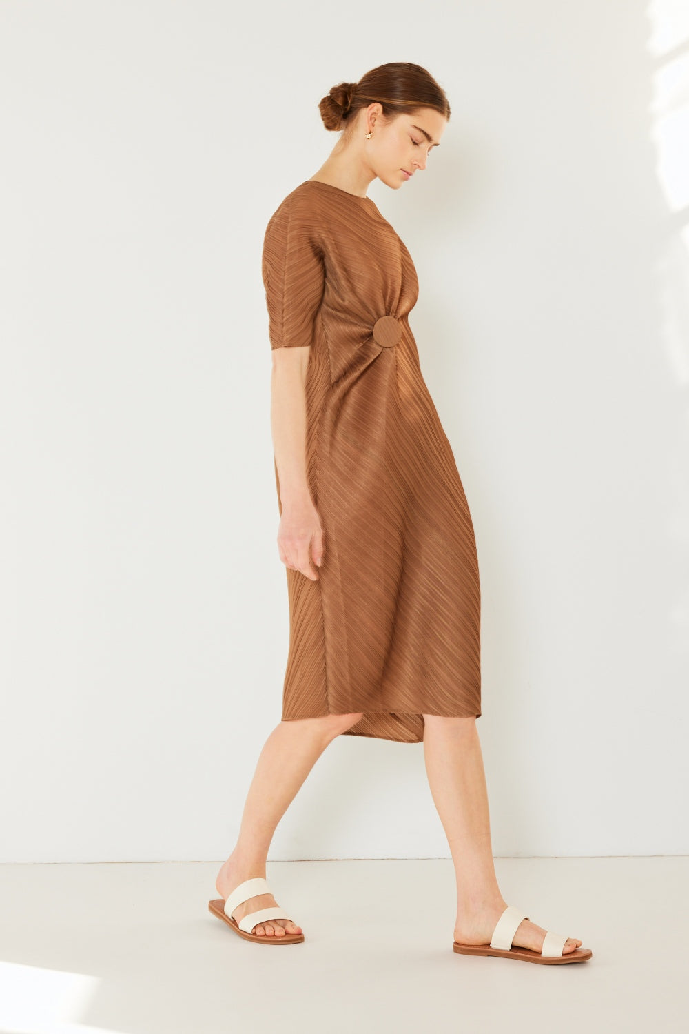 A woman stands against a plain backdrop wearing the stylish Marina West Swim Pleated Dolman Sleeve Dress, which features a knee-length, short-sleeved design in a textured brown fabric with an eye-catching circular detail at the waist. She completes her look with white sandals. This modern chic dress exudes versatility, making it perfect for various occasions.