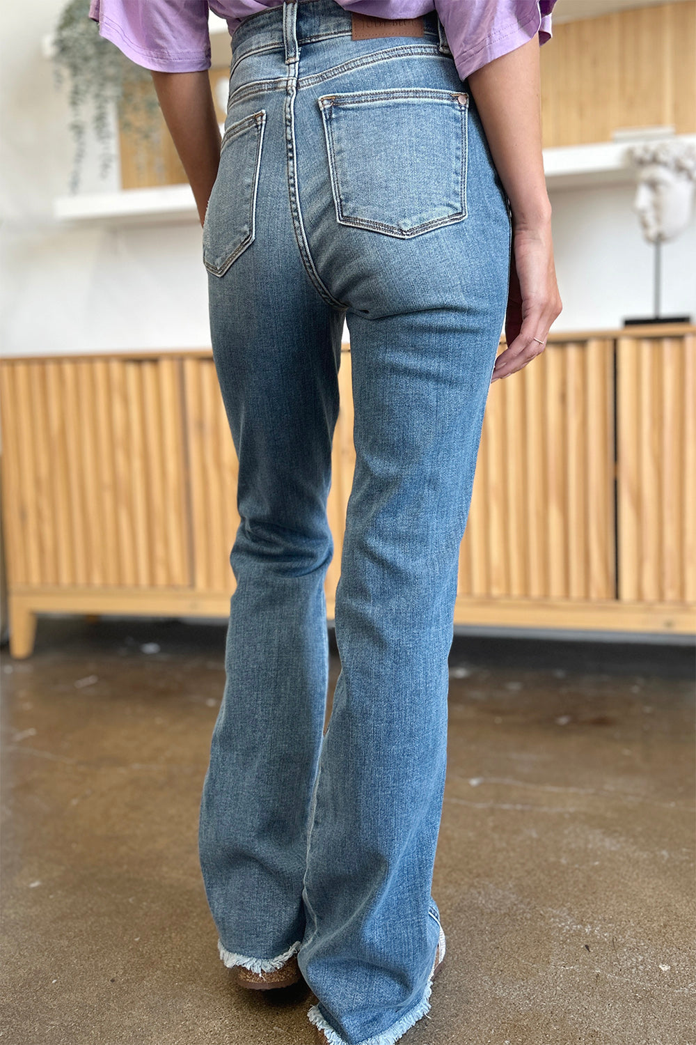 A person wearing Judy Blue Full Size Distressed Raw Hem Bootcut Jeans and a light purple top stands on a polished floor with a wooden cabinet in the background.