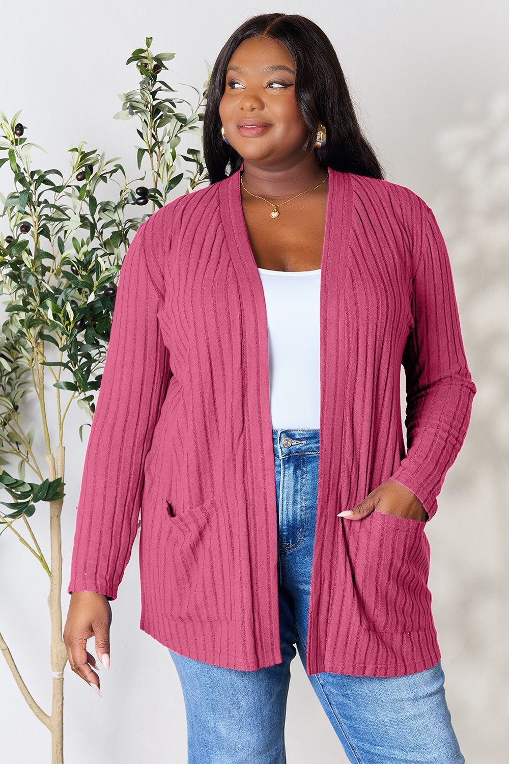 A woman in a Basic Bae Full Size Ribbed Open Front Cardigan with Pockets and jeans, made from stretchy material, stands in front of a plant, smiling slightly and looking to the side.