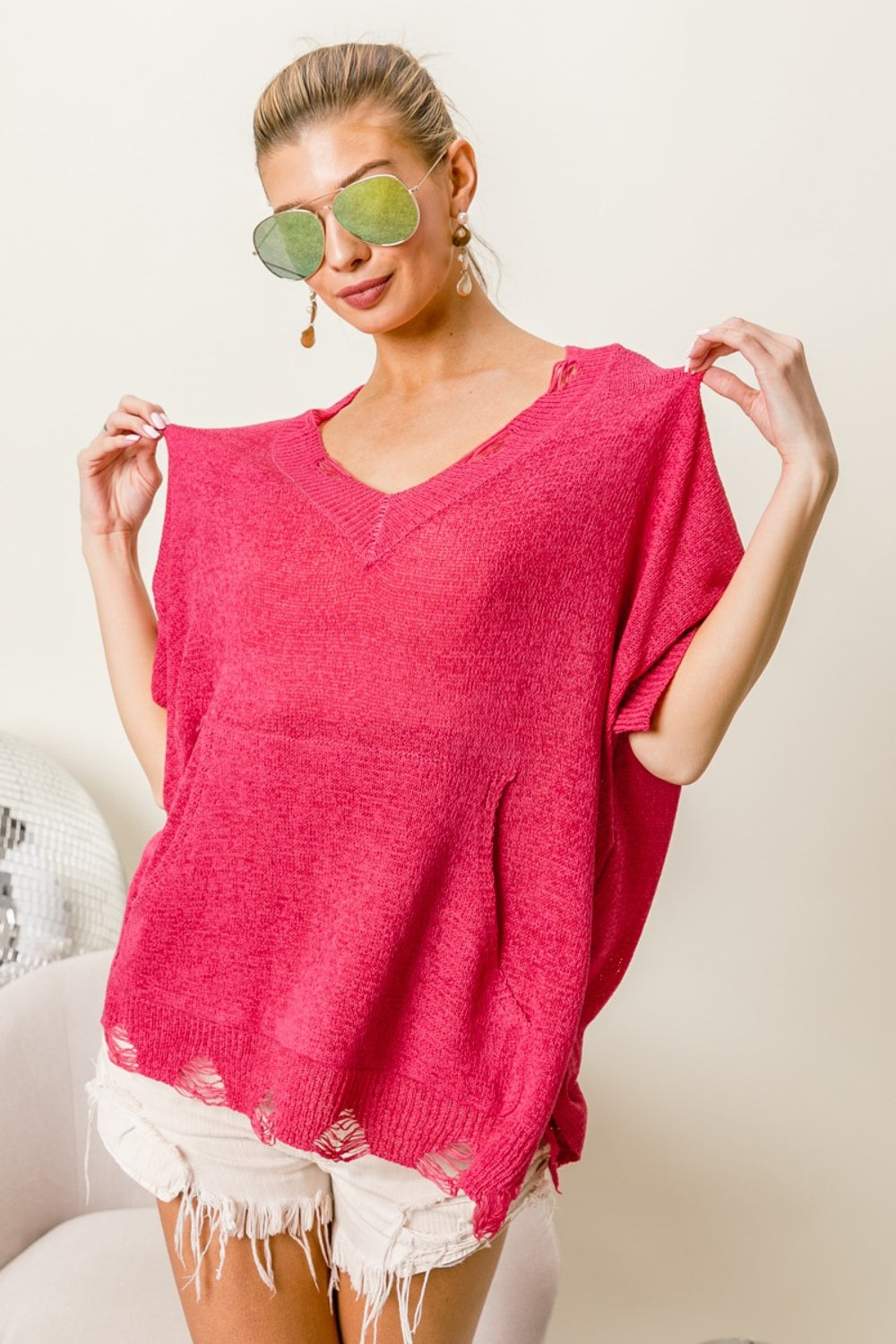 A woman in sunglasses, wearing a BiBi Distressed Hem V-Neck Slit Sweater in red and white shorts, smiles with her hands in her pockets, effortlessly showcasing her modern edge fashion.