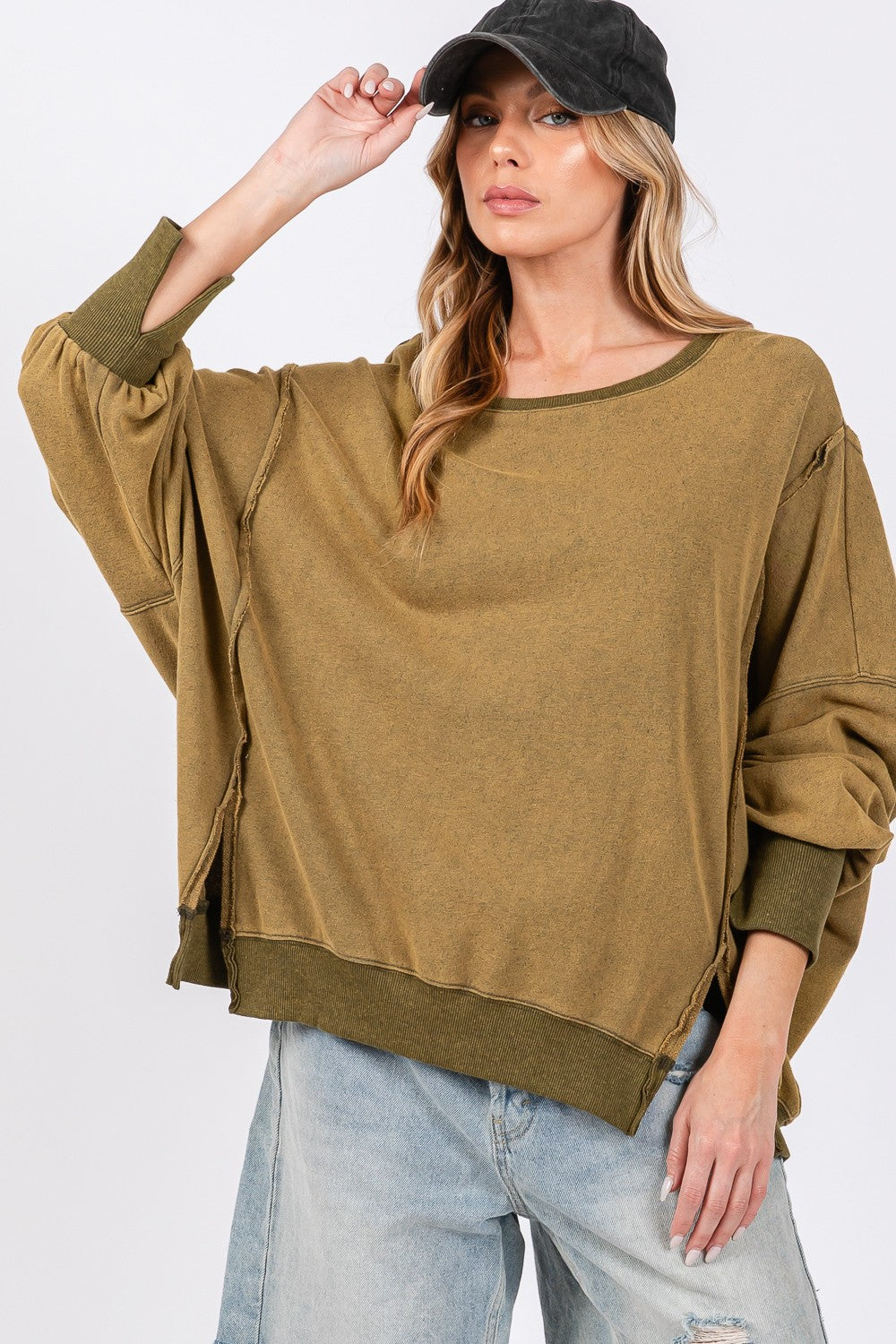 A person wearing a loose, oversized mineral wash SAGE + FIG Side Slit Sweatshirt in olive green, light blue jeans, and a black cap poses against a plain background.