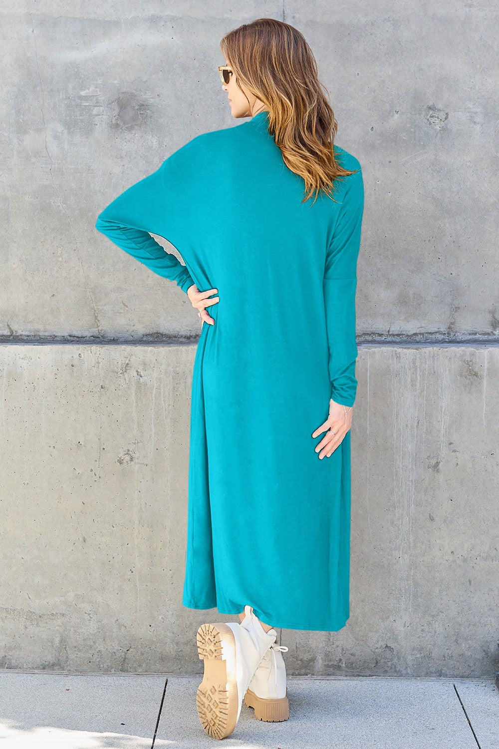 A woman stands against a concrete wall, wearing the Basic Bae Full Size Open Front Long Sleeve Cover Up made from rayon spandex, a white top, blue jeans in her perfect size, and brown shoes with her hands in her pockets. Machine wash cold for best results.