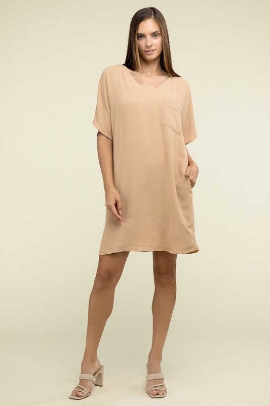 A woman wearing a loose light brown Woven Airflow V Neck T-Shirt Dress with Pockets stands against a plain background. Made from breathable fabric, this summer dress includes pockets where her hands rest as she smiles slightly.