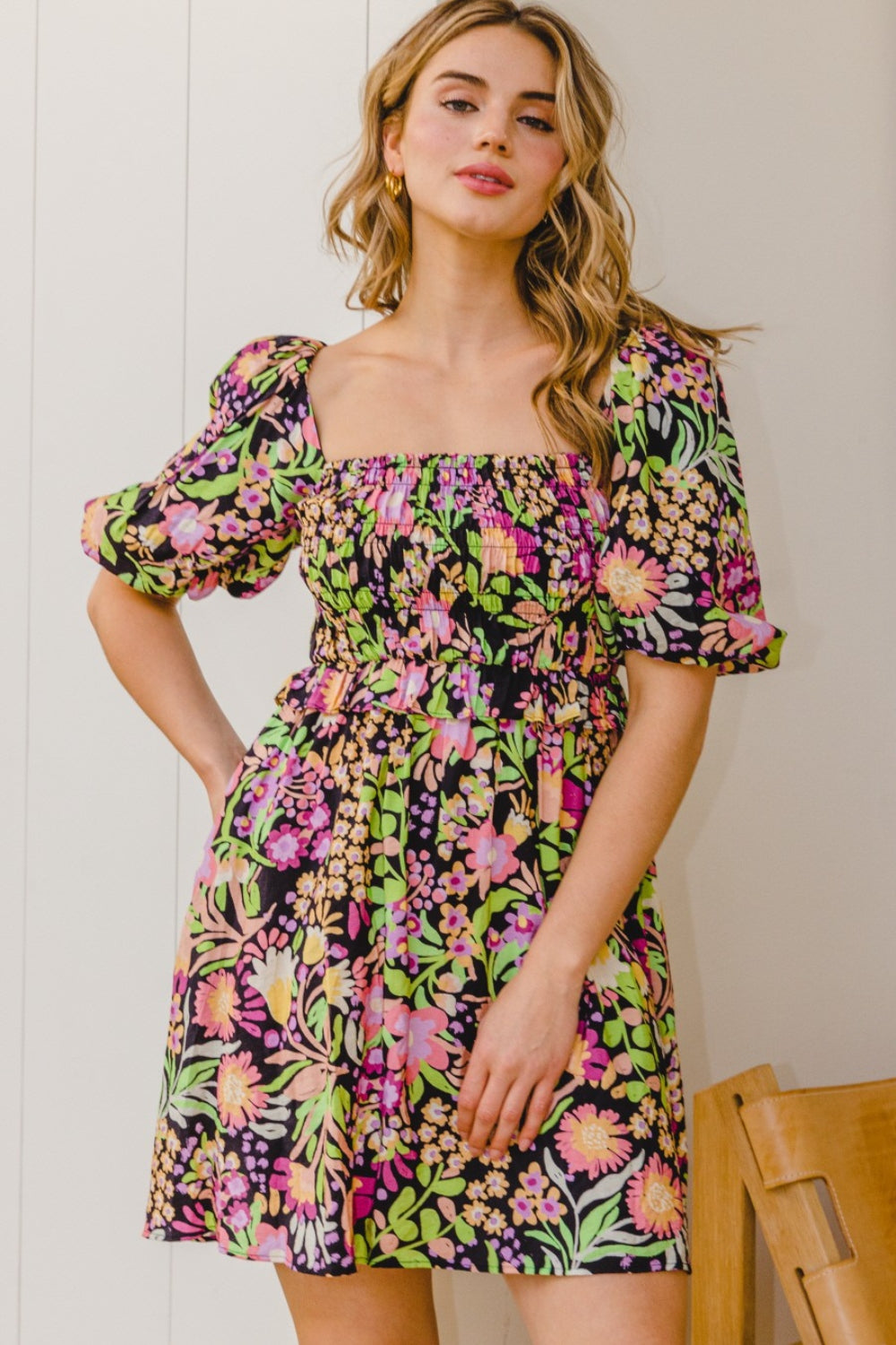 A person stands wearing the ODDI Full Size Floral Tie-Back Mini Dress, a feminine and flirty black floral design with puffy sleeves, perfect for a summer wedding.