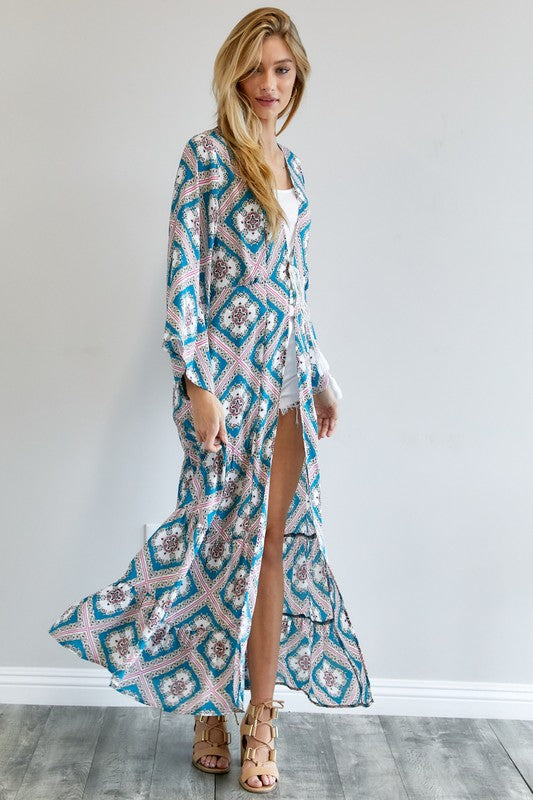 A person wearing a Printed Long Sleeve Loose Kimono with a bohemian blue and pink print over a white outfit stands in a room with a gray floor and white wall.