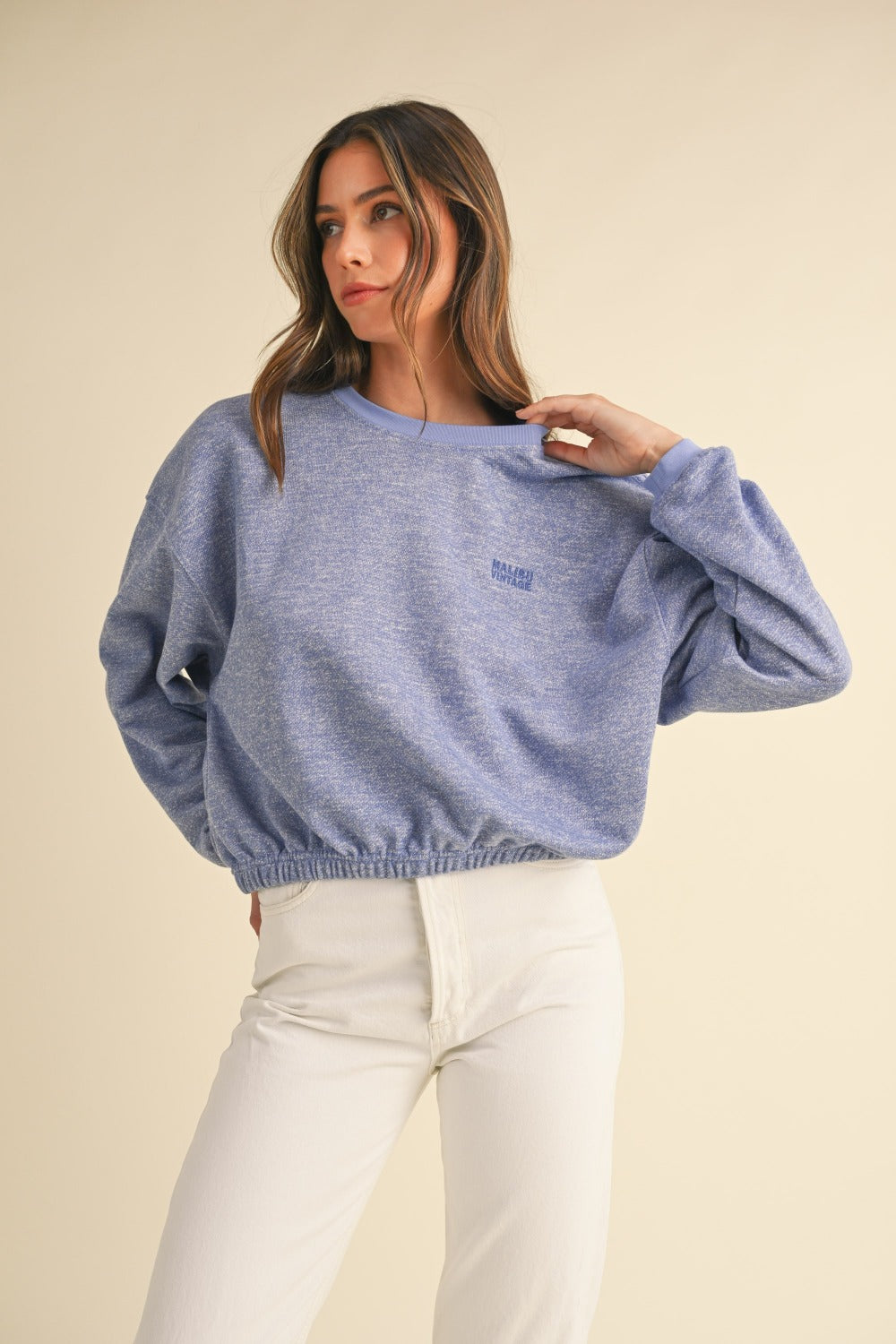 A person wearing a Mable Round Neck Letter Embroidery Crop Sweatshirt in blue, paired with white pants, stands against a plain background, touching the collar of the sweatshirt with one hand.