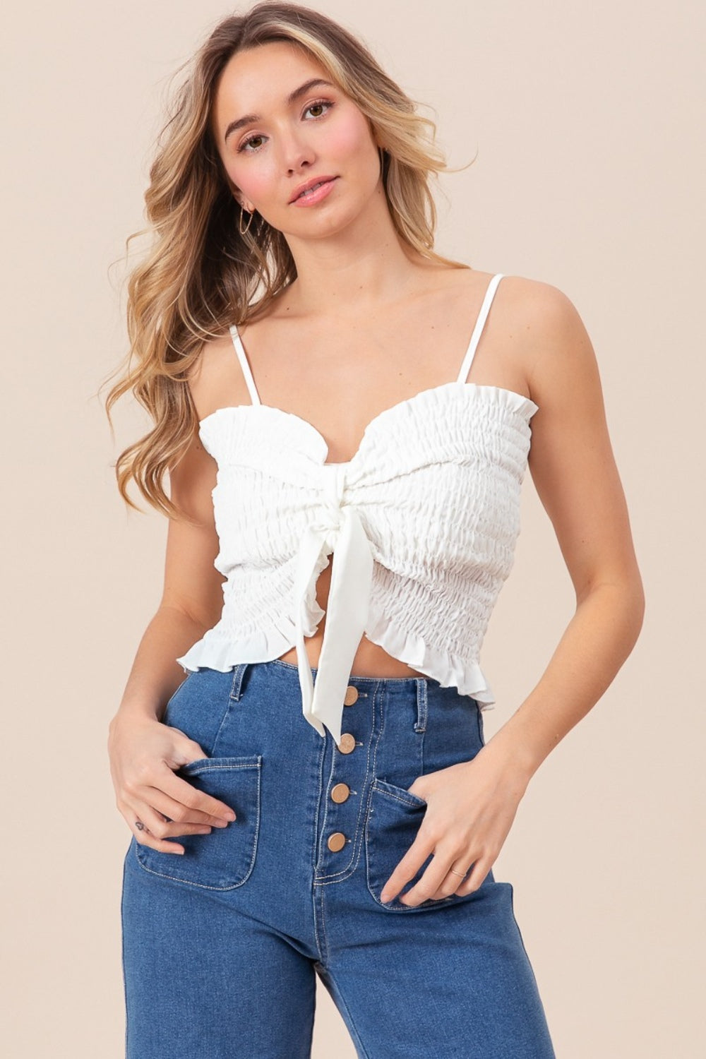 A woman wearing the BiBi Ruffled Smocked Ribbon Detail Cami in white, paired with blue high-waisted jeans, is posing against a neutral background.