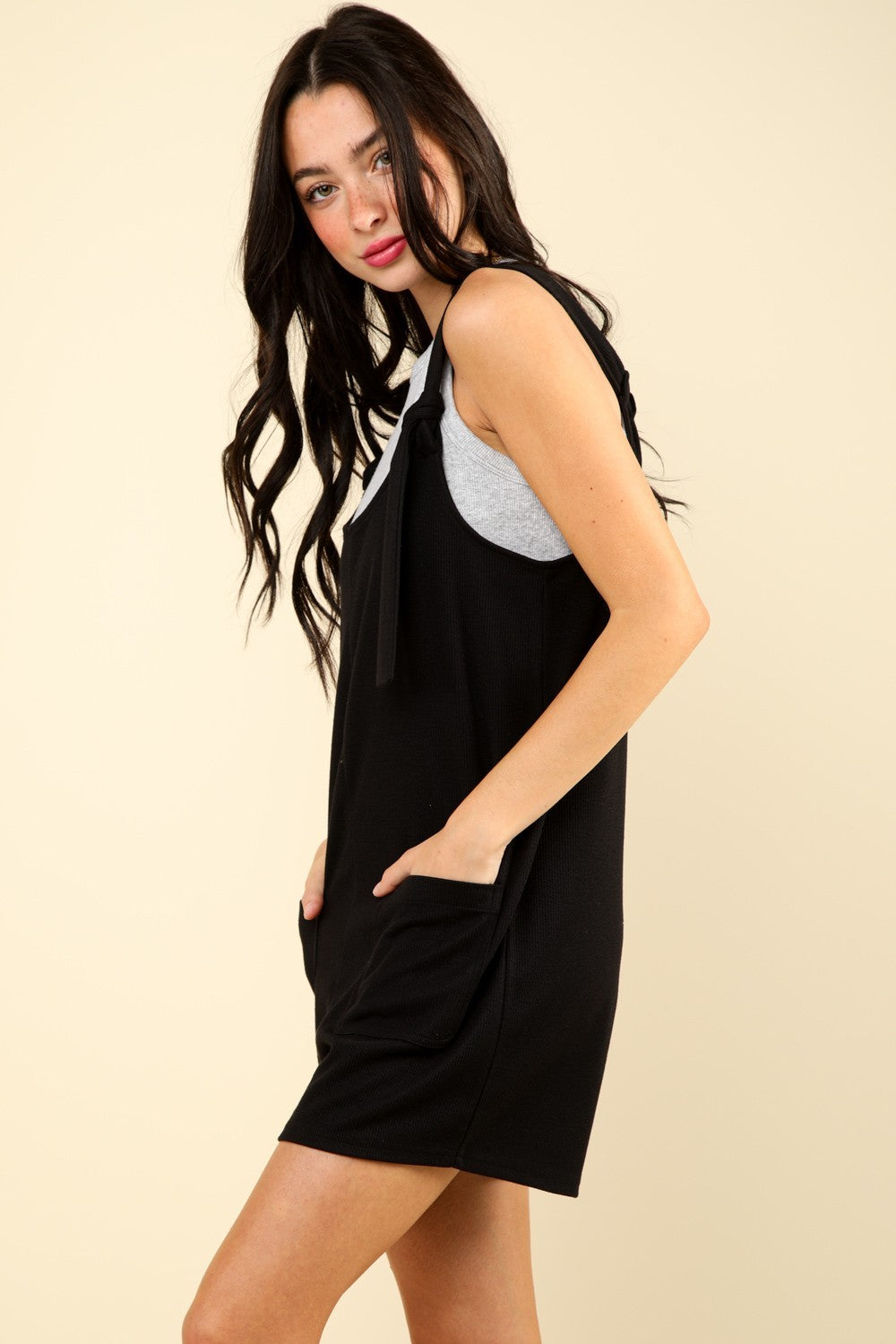 A person with long dark hair stands against a plain background, wearing the VERY J Tie Shoulder Front Pocket Romper in gray, which features tied shoulder straps and a casual knit style, smiling slightly.