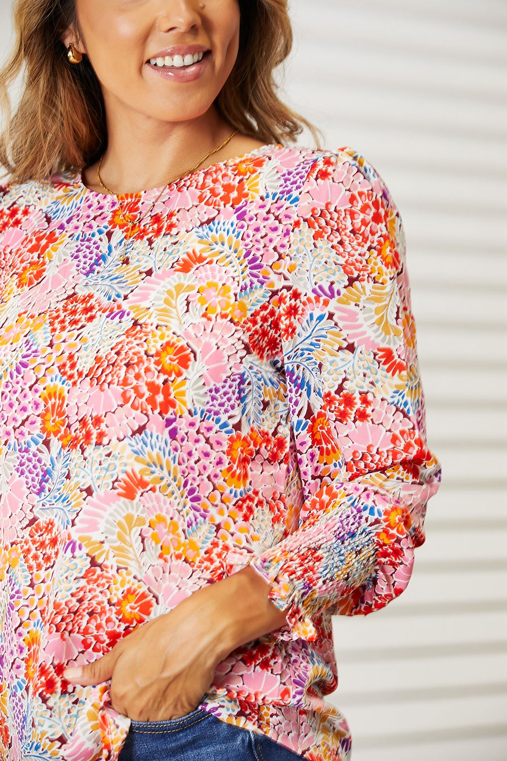 A smiling woman wearing a Double Take Floral Print Long Puff Sleeve Blouse and blue jeans stands in a brightly lit room.