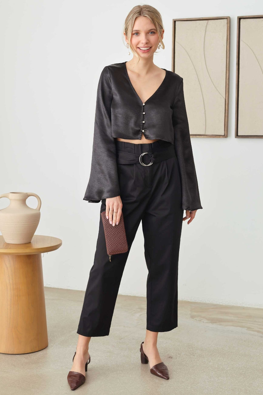 A person in a black long-sleeve Mustard Seed Satin Crop Top with rhinestone button closures and pants, holding a clutch, stands near a wooden table with a vase. Two framed artworks elegantly hang on the wall behind.