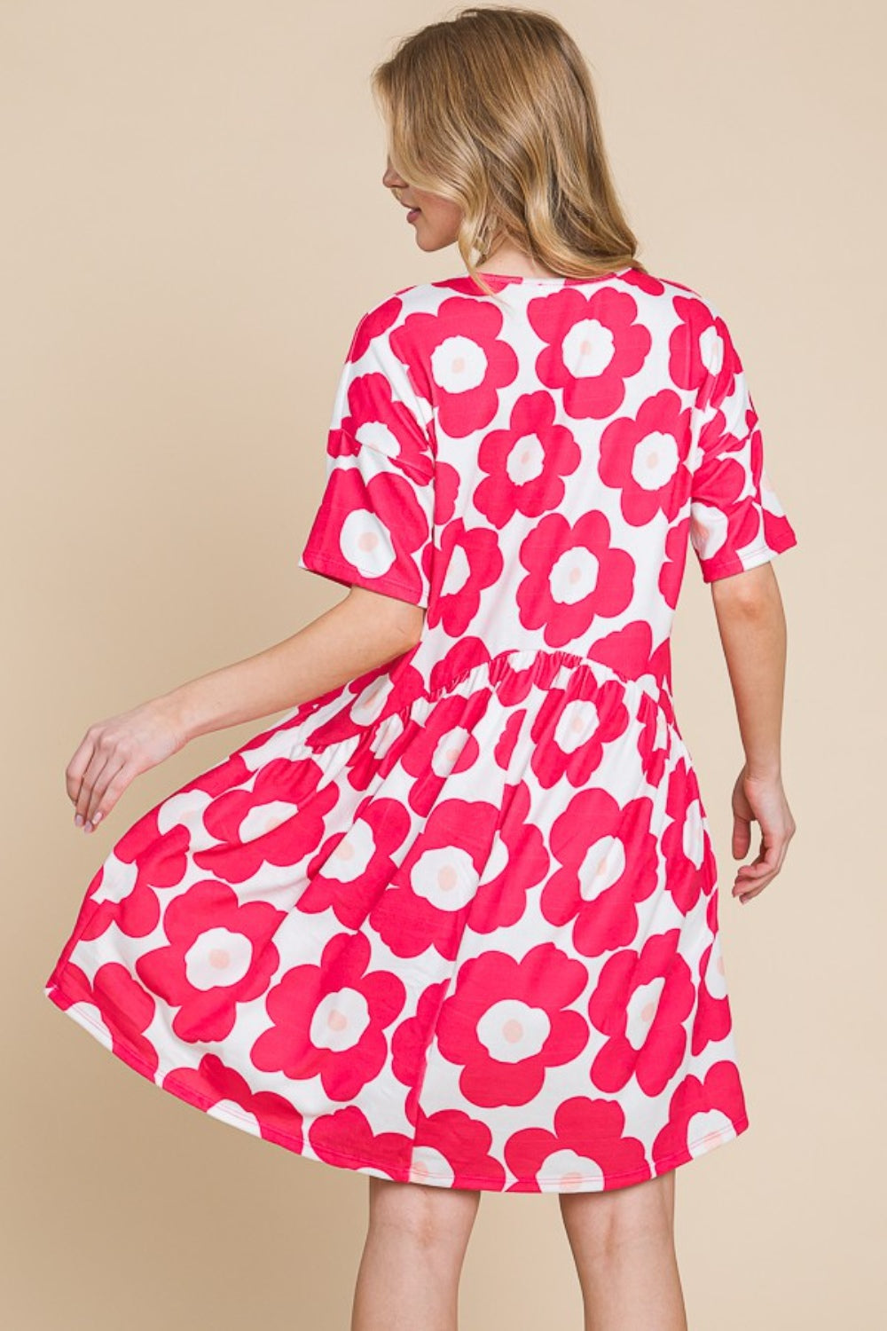 A woman gracefully stands against a beige background, wearing the BOMBOM Flower Print Ruched Dress adorned with large pink blooms. The stretchy fabric of the dress complements her form, effortlessly enhancing its elegant silhouette.