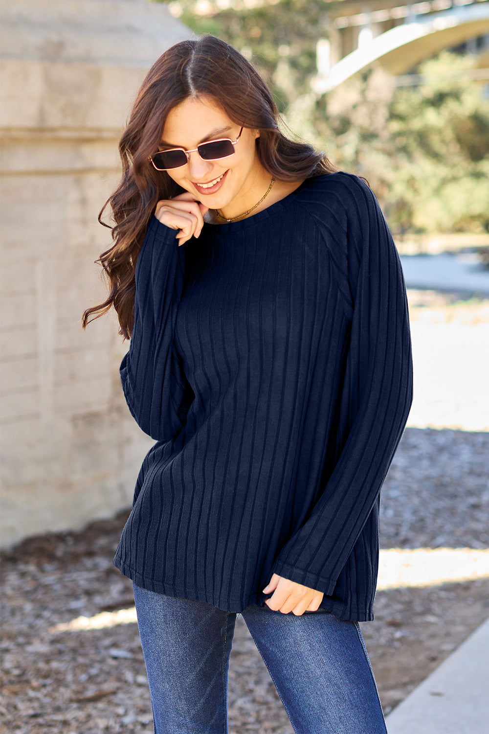 A woman with long brown hair wearing sunglasses and a Basic Bae full-size, slightly stretchy ribbed round neck long sleeve knit top in red, paired with blue jeans, is standing outdoors.