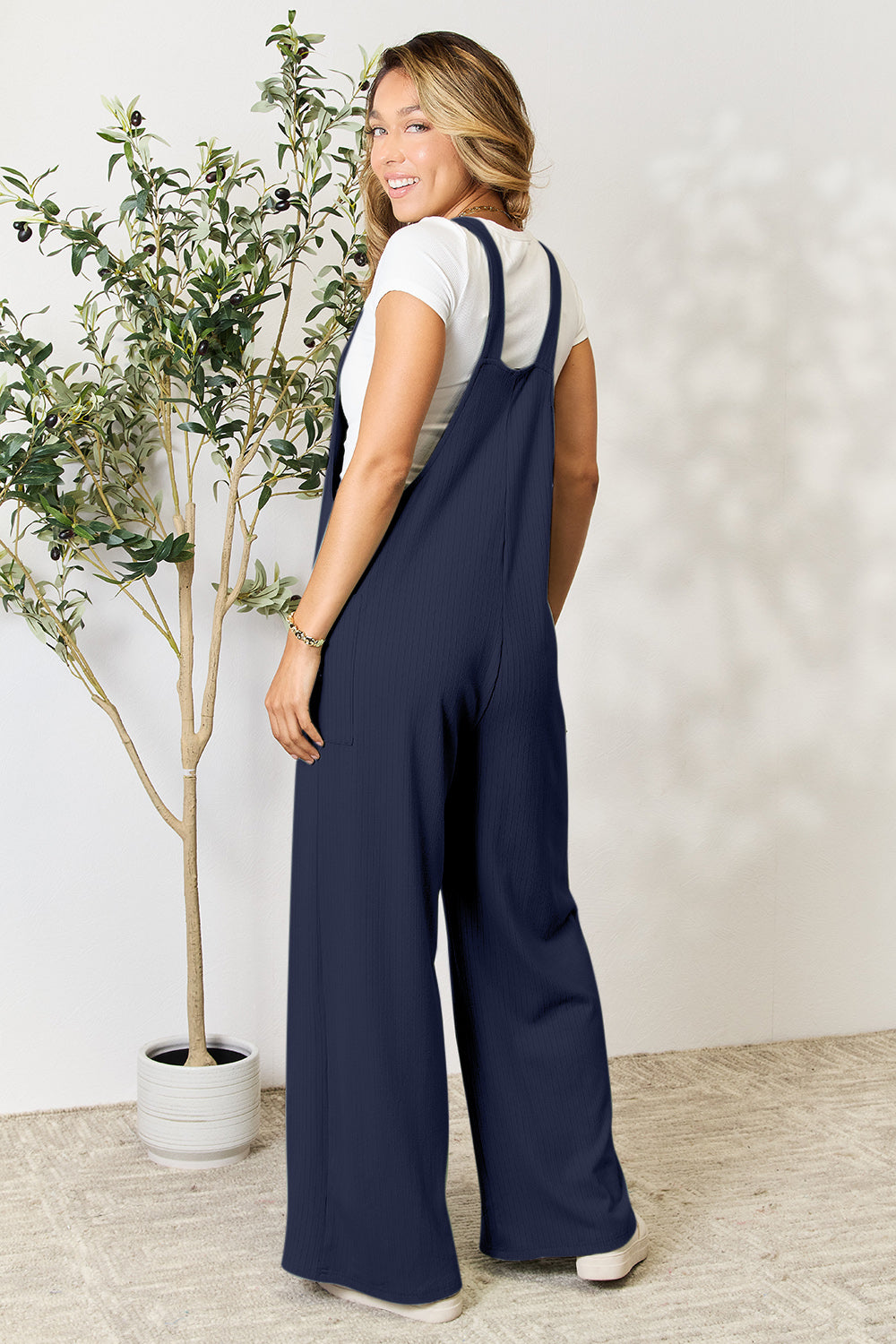 A smiling person wearing the "Double Take Full Size Wide Strap Overall with Pockets," known for its basic style and size-inclusive fit, stands indoors next to a potted plant.