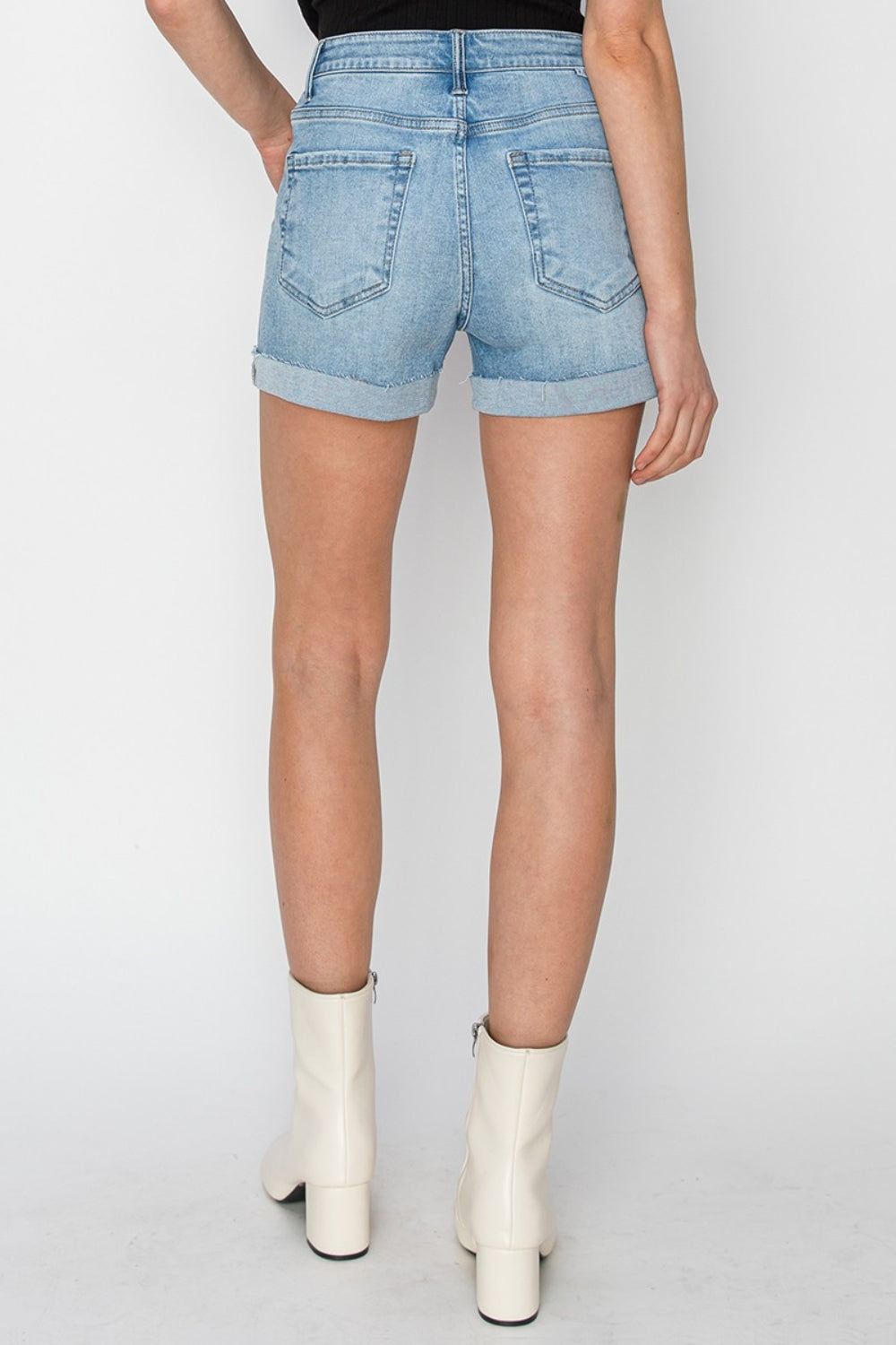 A person wearing RISEN distressed mid-rise waist denim shorts in light blue paired with white ankle boots and a black top, standing against a plain background—a staple for any summer wardrobe.