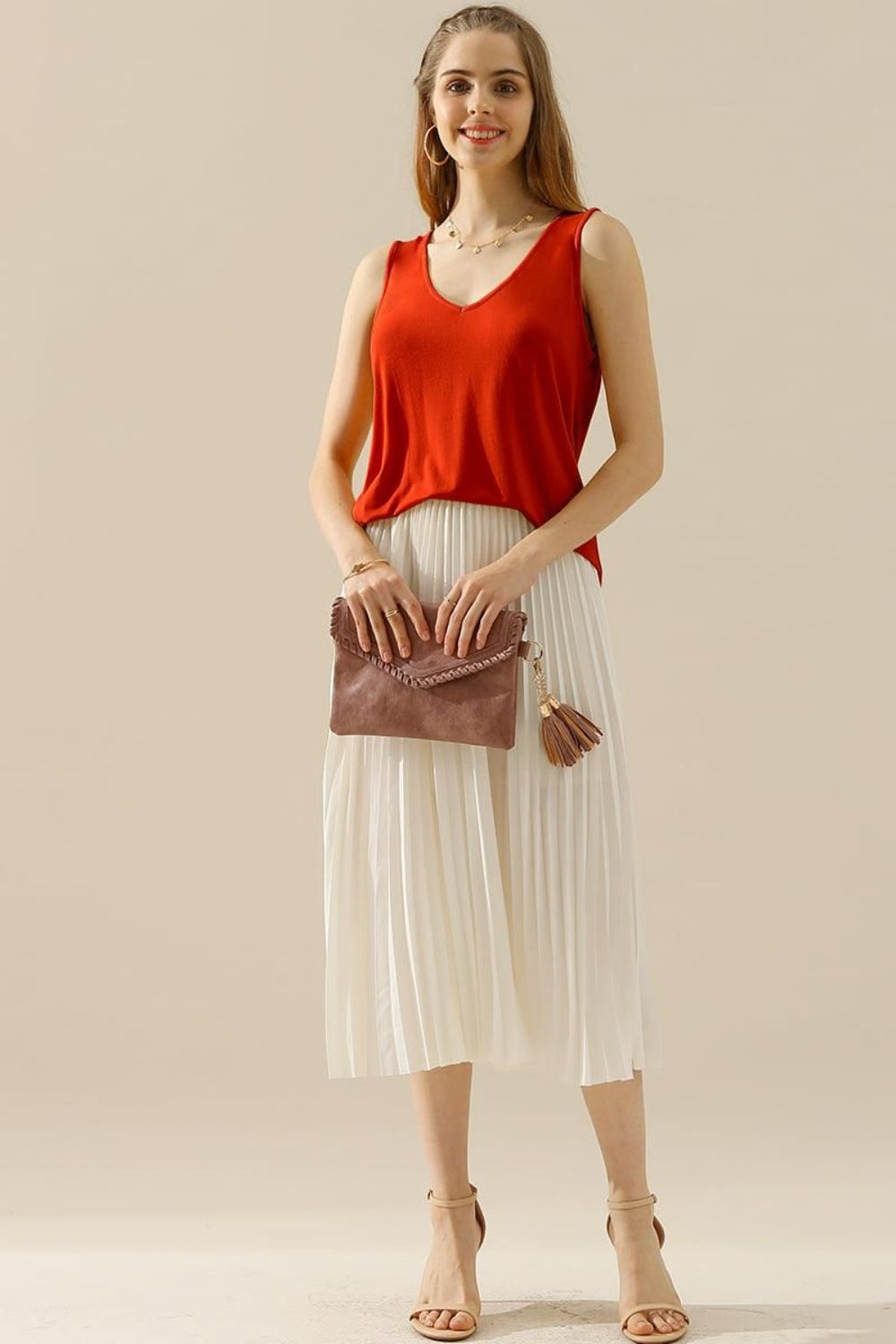 A woman in a stylish and versatile Ninexis Full Size V-Neck Curved Hem Tank and white pleated skirt poses while holding her handbag.