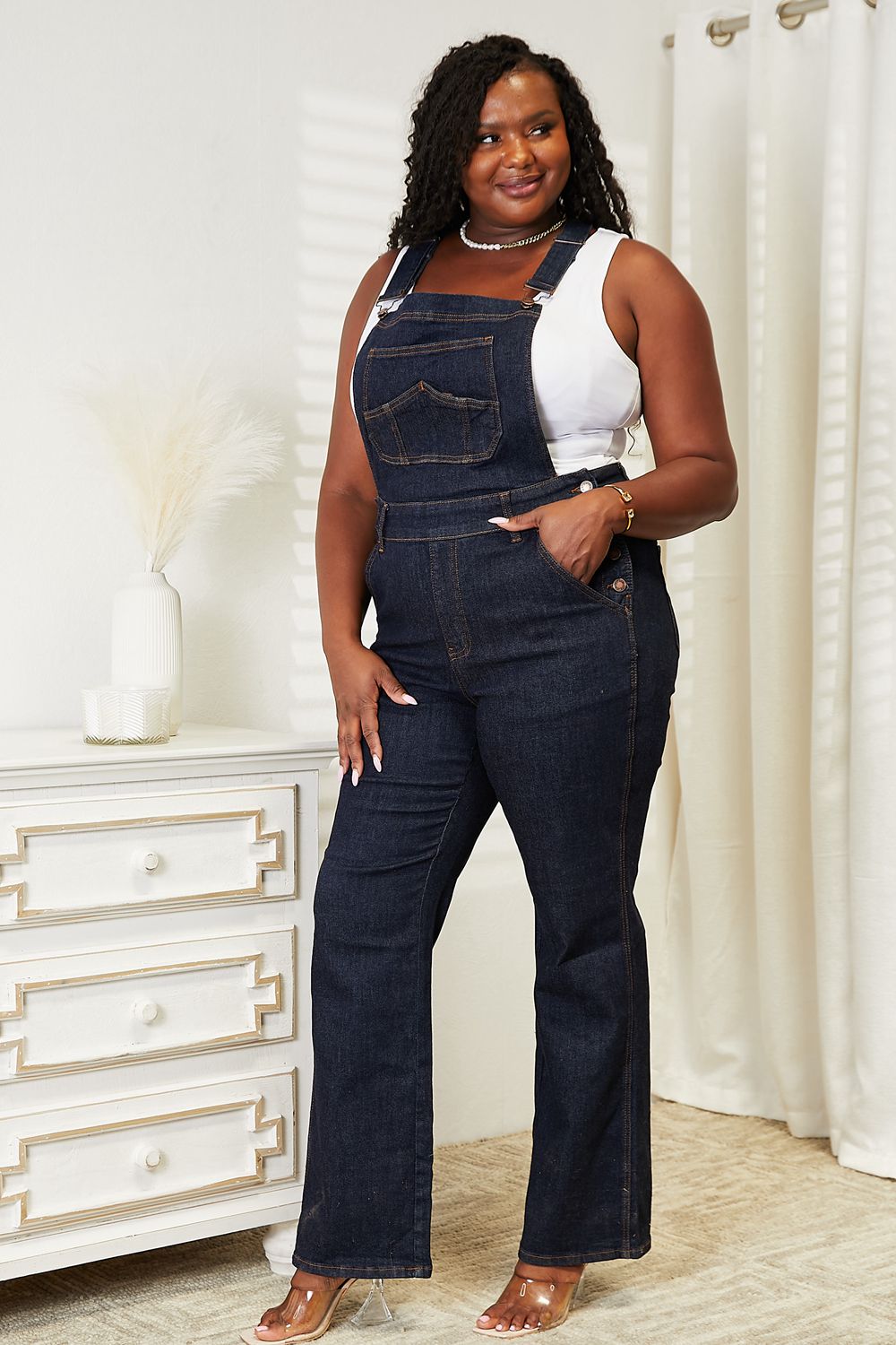 A person stands indoors wearing Judy Blue Full Size High Waist Classic Denim Overalls and a white tank top, with one hand in the pocket and the other resting on a white chest of drawers, next to a vase of dried pampas grass.