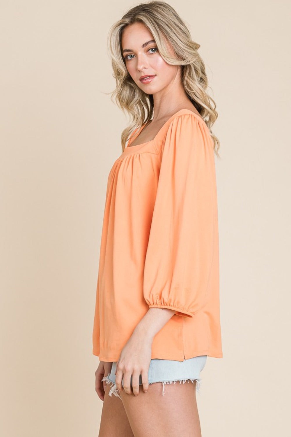 A person with long blonde hair is smiling and wearing the trendy Culture Code Square Neck Puff Sleeve Top in a light orange hue, paired with light denim shorts, showcasing a versatile fashion piece against a beige background.