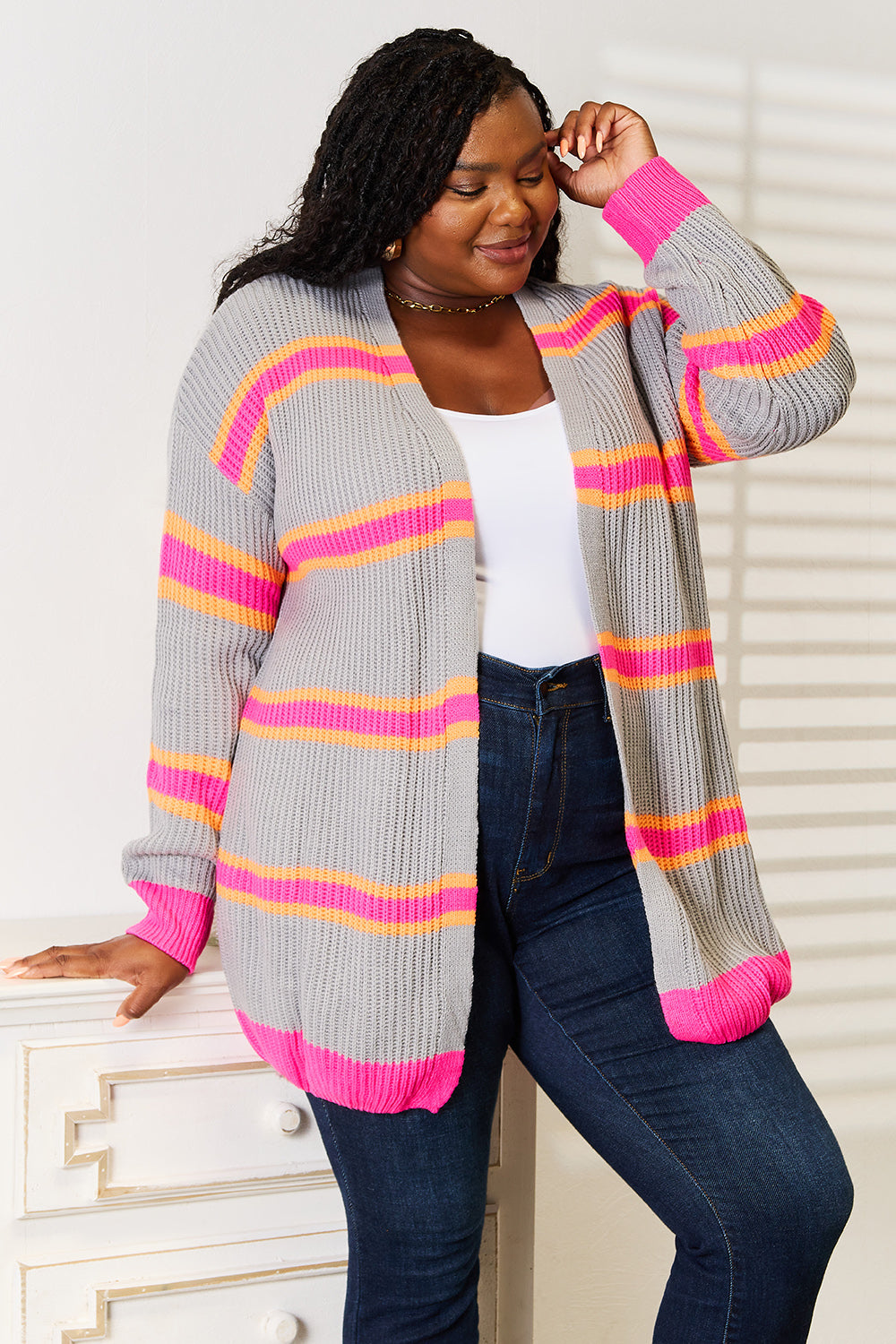 A woman stands against a white wall, smiling with one hand on her hip. She wears the Woven Right Ribbed Long Sleeve Cardigan, featuring pink and orange stripes, over a white top and dark jeans—an excellent choice for versatile layering.