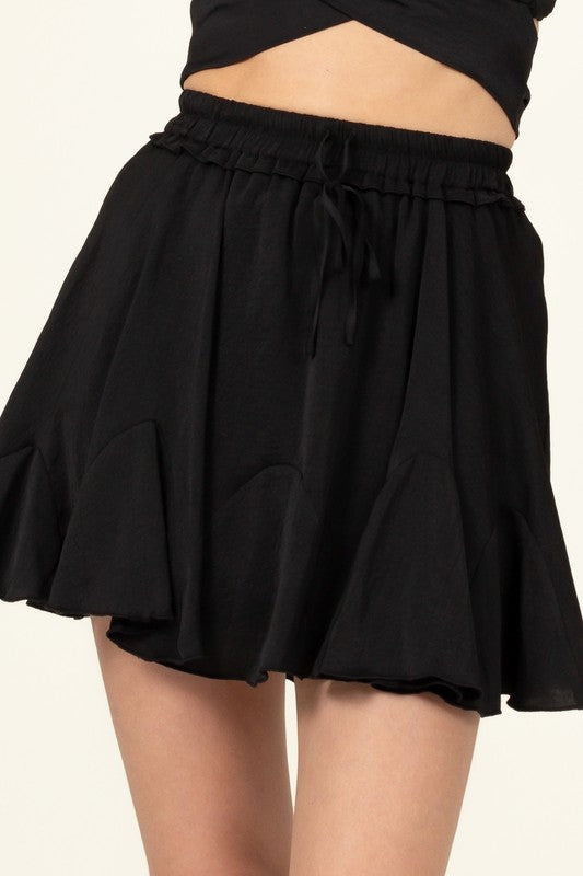 A person is partially visible in the image wearing the Not Your Girl Drawstring Ruffled Mini Skirt, featuring a bright pink flared silhouette with an elastic waistband and drawstring, paired with a matching top.