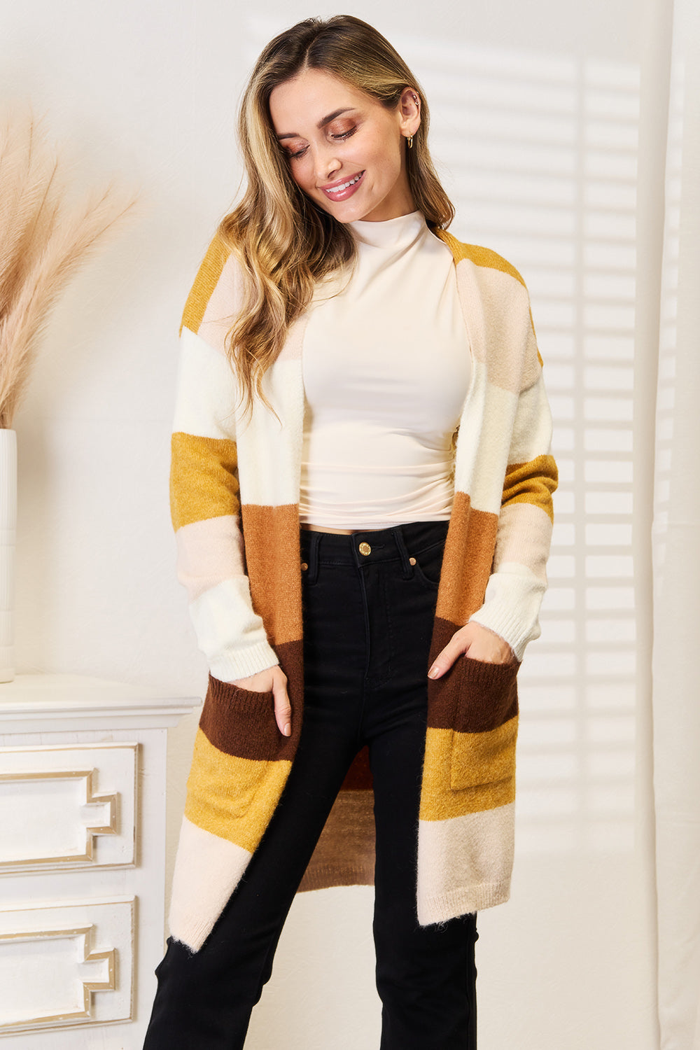 A woman wearing a cozy Woven Right Color Block Dropped Shoulder Cardigan, white turtleneck, and black pants stands indoors with her hands in her pockets.