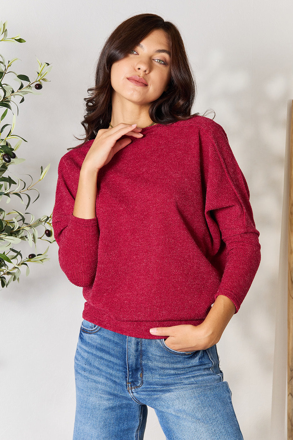 Person wearing a BOMBOM Drop Shoulder Long Sleeve Blouse in red, paired with blue jeans, standing with one hand in their pocket and the other touching their neck, in front of a white wall with a potted plant on the side.