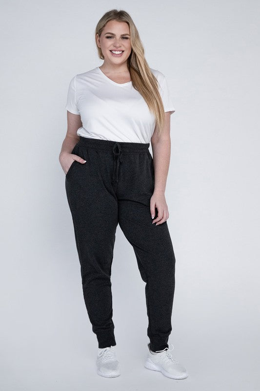 A person stands against a white background, wearing a white t-shirt, grey Plus-Size Jogger Pants with an adjustable waistband, and white sneakers. They have long blonde hair and are posing with one hand on their hip.