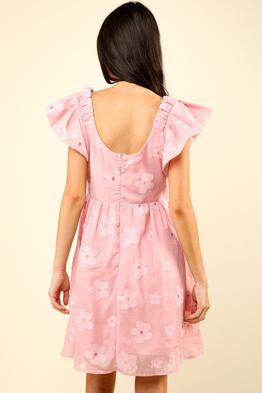 Woman in a VERY J Flower Embroidered Organza Mini Dress with flutter sleeves stands against a light background, resting one hand on the skirt and the other slightly bent. She is wearing strappy sandals.