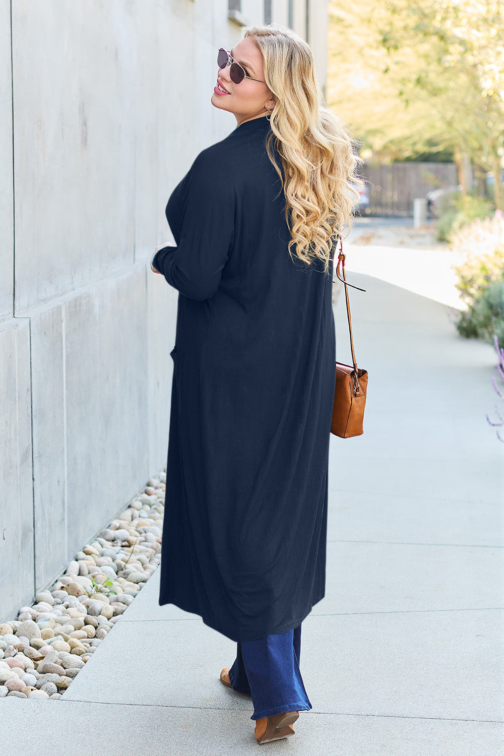 A woman stands against a concrete wall, wearing the Basic Bae Full Size Open Front Long Sleeve Cover Up made from rayon spandex, a white top, blue jeans in her perfect size, and brown shoes with her hands in her pockets. Machine wash cold for best results.