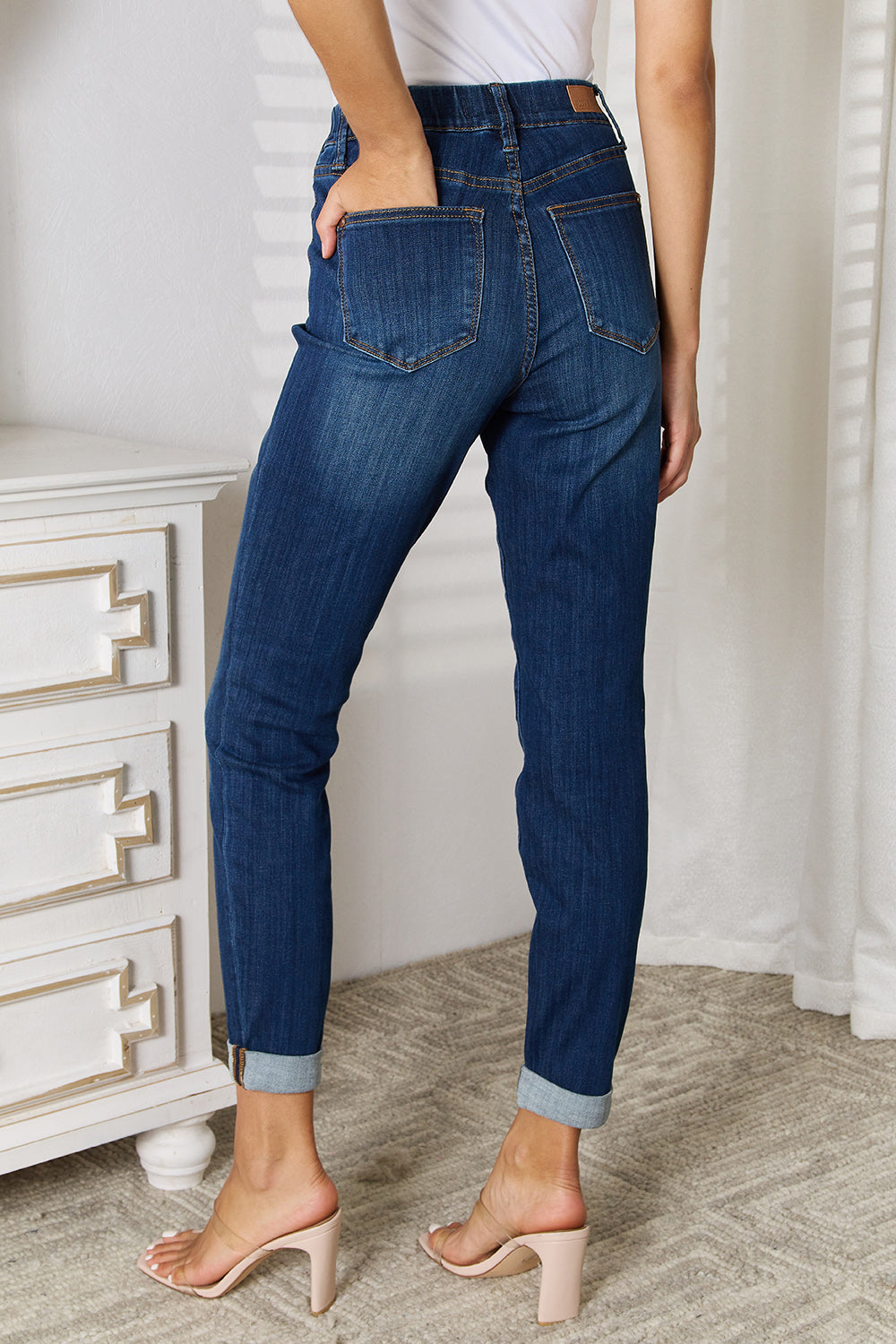 A person wearing Judy Blue Full Size Skinny Cropped Jeans with rolled-up cuffs and clear heels stands near a white dresser adorned with a decorative vase for a modern look.