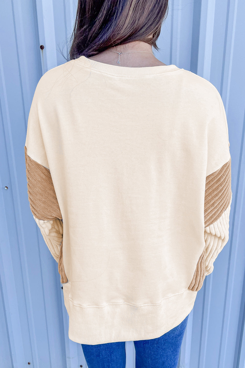 A person with long hair is wearing an Apricot Corduroy Patchwork Drop Shoulder Long Sleeve Sweatshirt, standing with their back to the camera against a light blue corrugated metal wall.