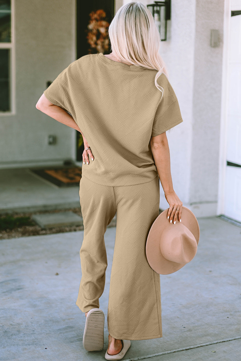 A woman with long hair smiles while standing outside a building, dressed in the Double Take Full Size Texture Short Sleeve Top and Pants Set, featuring a matching beige two-piece ensemble. She wears slides and holds a hat in her right hand. Her outfit, slightly stretchy for added comfort, is perfect for machine wash cold.