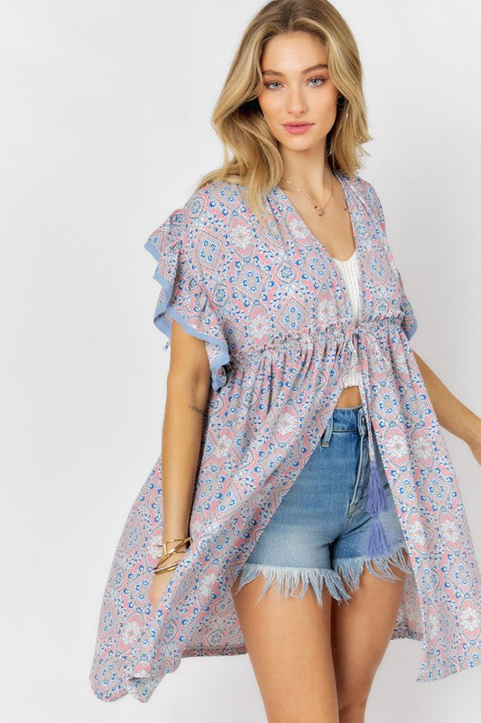 A woman stands against a white background wearing the Printed Short Sleeve Ruffle Kimono over a white top and denim shorts, effortlessly blending style and comfort.