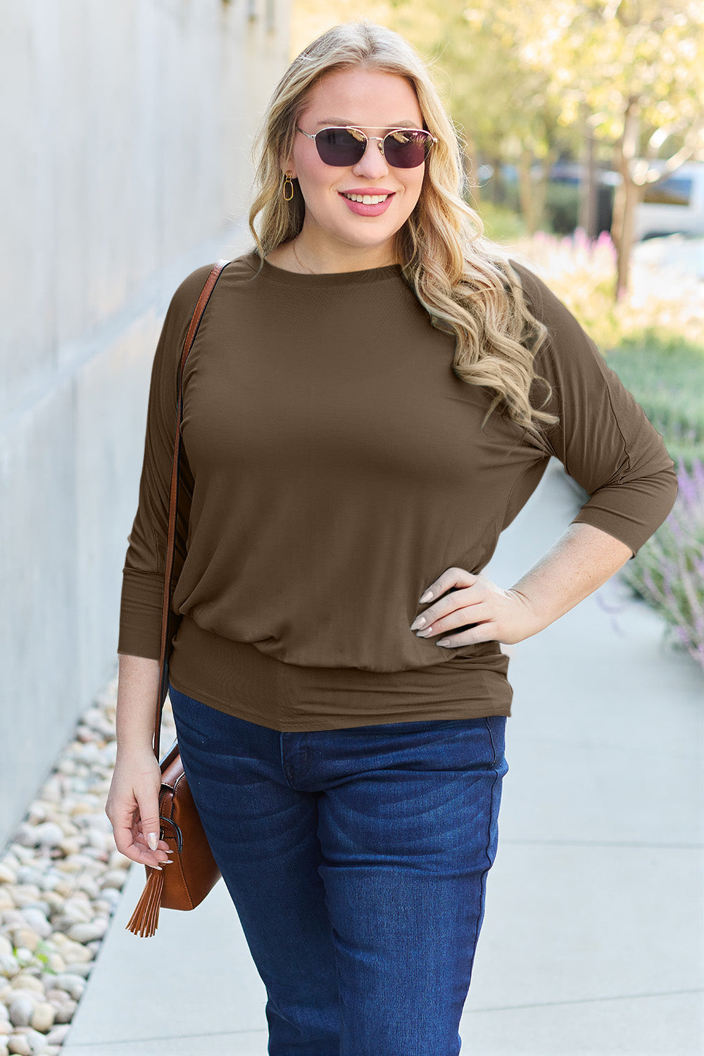 A woman in a slightly stretchy, brown Basic Bae Full Size Round Neck Batwing Sleeve top and blue jeans stands against a concrete wall, holding a brown handbag.