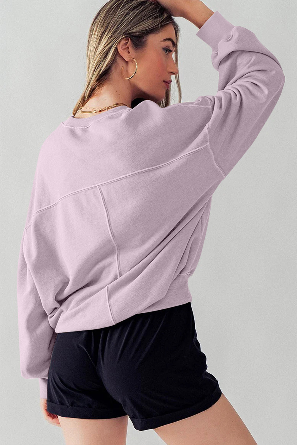 A woman facing away wears the Orchid Petal Exposed Seam Batwing Sleeve Drop Shoulder Sweatshirt and black shorts. She poses with her right arm raised, resting on her head.