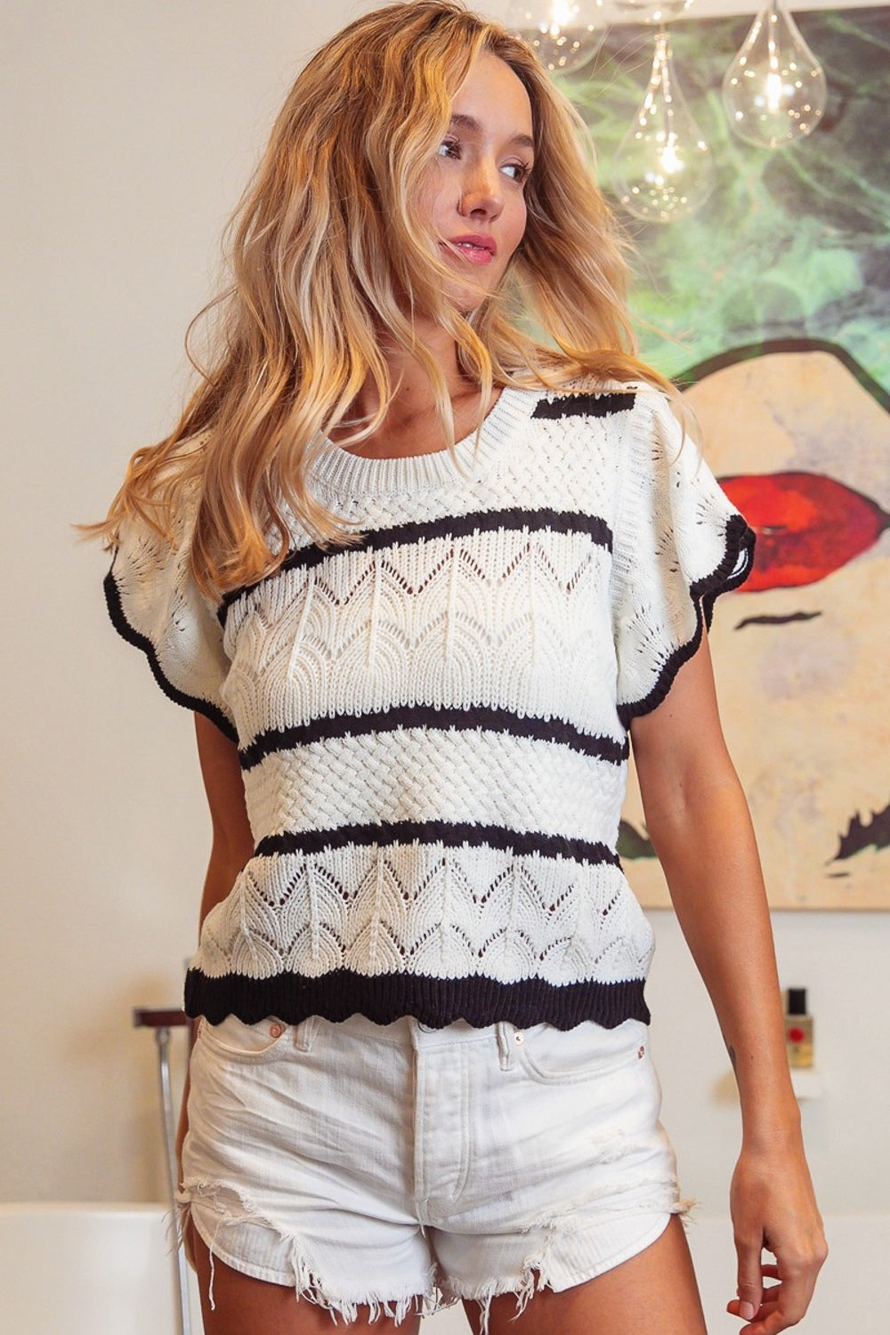 A woman with long blonde hair wearing a BiBi Pointelle Contrast Striped Short Sleeve Knit Top and white shorts smiles in a brightly lit room. A colorful painting is visible in the background, adding a touch of vibrancy to her sophisticated, stylish wardrobe.