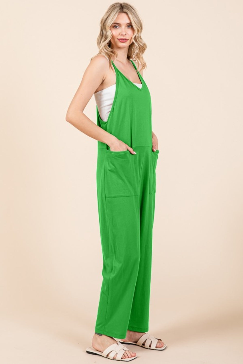 A person stands against a plain background wearing the Culture Code Full Size Sleeveless Jumpsuit with Pockets in green, paired with white sandals, showcasing its practical pockets with hands tucked comfortably inside.