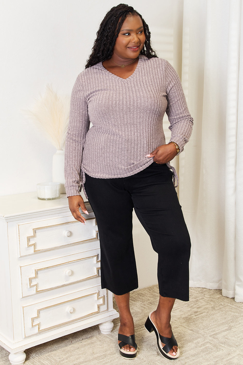 A woman stands indoors against a neutral background, wearing the "Double Take Drawstring Ribbed Long Sleeve T-Shirt" in light purple paired with dark pants. She is smiling and posing with one hand on her hip.