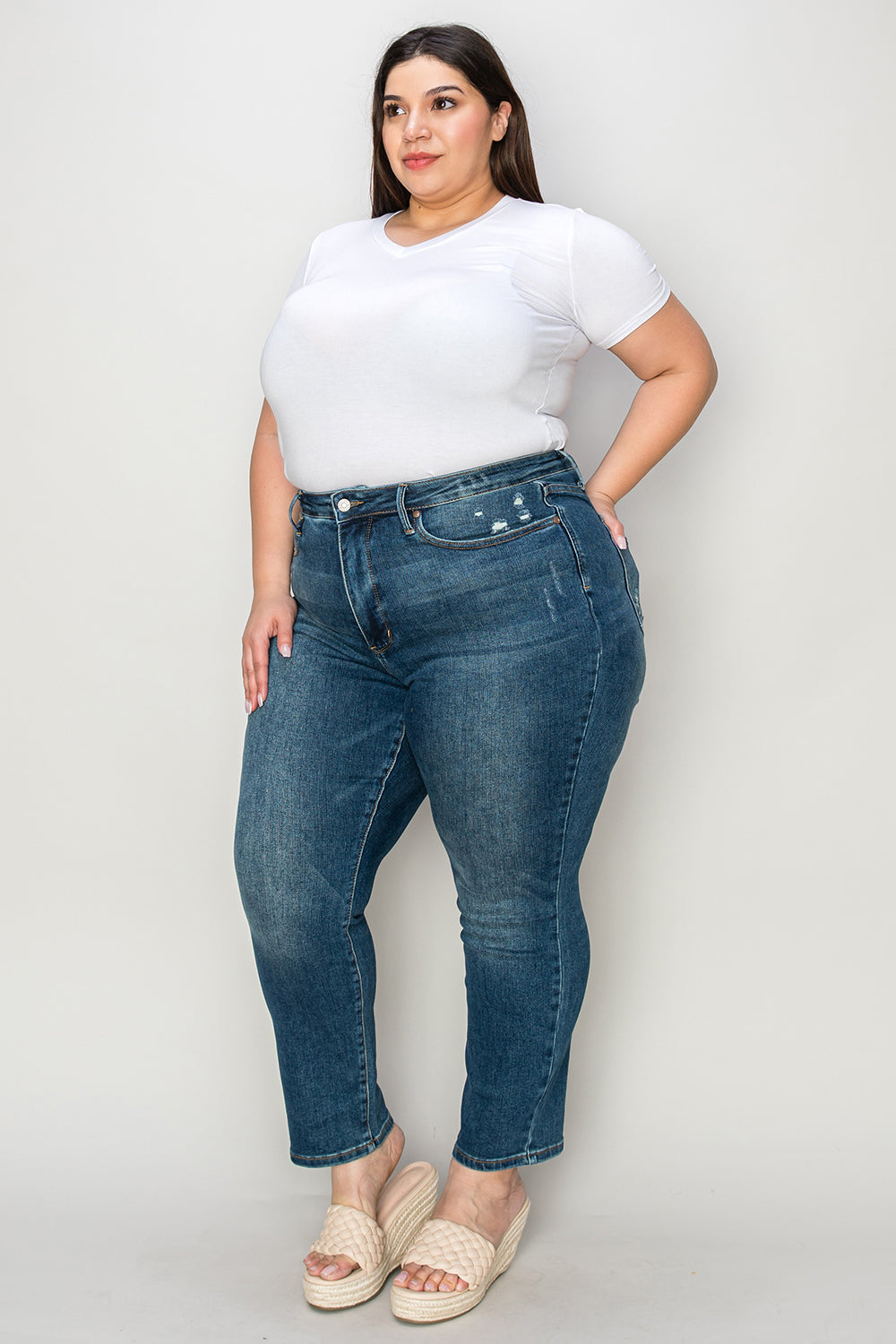 Person wearing Judy Blue Full Size Tummy Control High Waist Slim Jeans in blue and a white shirt, with their hands in their pockets. They are standing indoors on a polished concrete floor with a wooden cabinet and plant in the background.