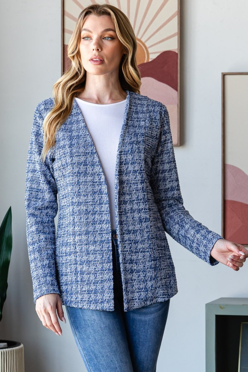 A woman with wavy blonde hair wears the Heimish Full Size Houndstooth Tweed Open Front Blazer in blue, paired with a white top and blue jeans. She stands in front of a wall adorned with framed art.