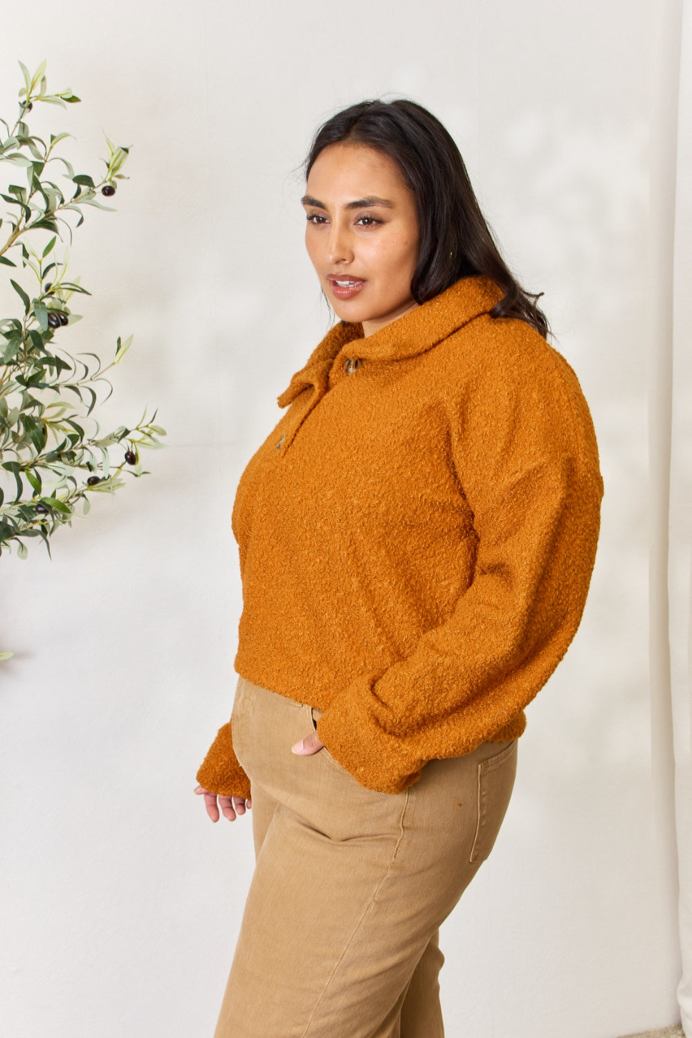 A person wears a brown textured Culture Code Full Size Half Button Turtleneck Sweatshirt made from breathable fabrics and tan pants, standing next to a leafy plant against a light background.