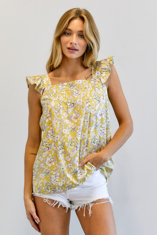 A woman in a vibrant summer wardrobe stands against a plain background, wearing the Floral Printed Ruffle Sleeveless Top paired with white shorts, with one hand casually tucked in her pocket.