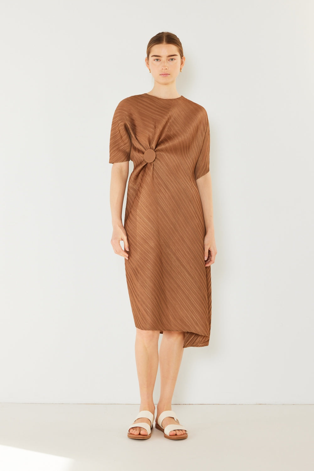 A woman stands against a plain backdrop wearing the stylish Marina West Swim Pleated Dolman Sleeve Dress, which features a knee-length, short-sleeved design in a textured brown fabric with an eye-catching circular detail at the waist. She completes her look with white sandals. This modern chic dress exudes versatility, making it perfect for various occasions.