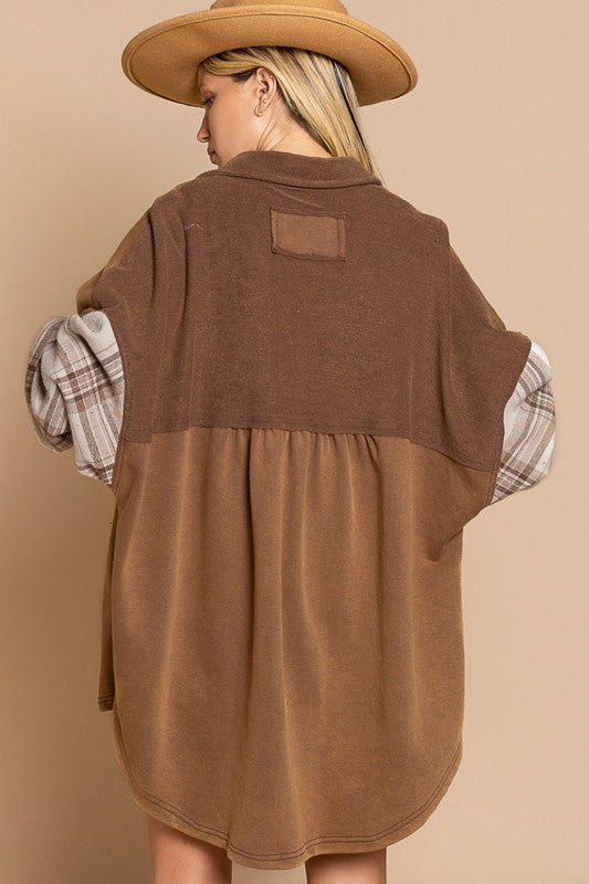 A person wearing the Long Sleeve With Plaid Detail Sleeve Shacket in brown, featuring plaid details on the sleeves and a matching pocket, paired with a white top underneath and a wide-brimmed hat—perfect for transitional weather.